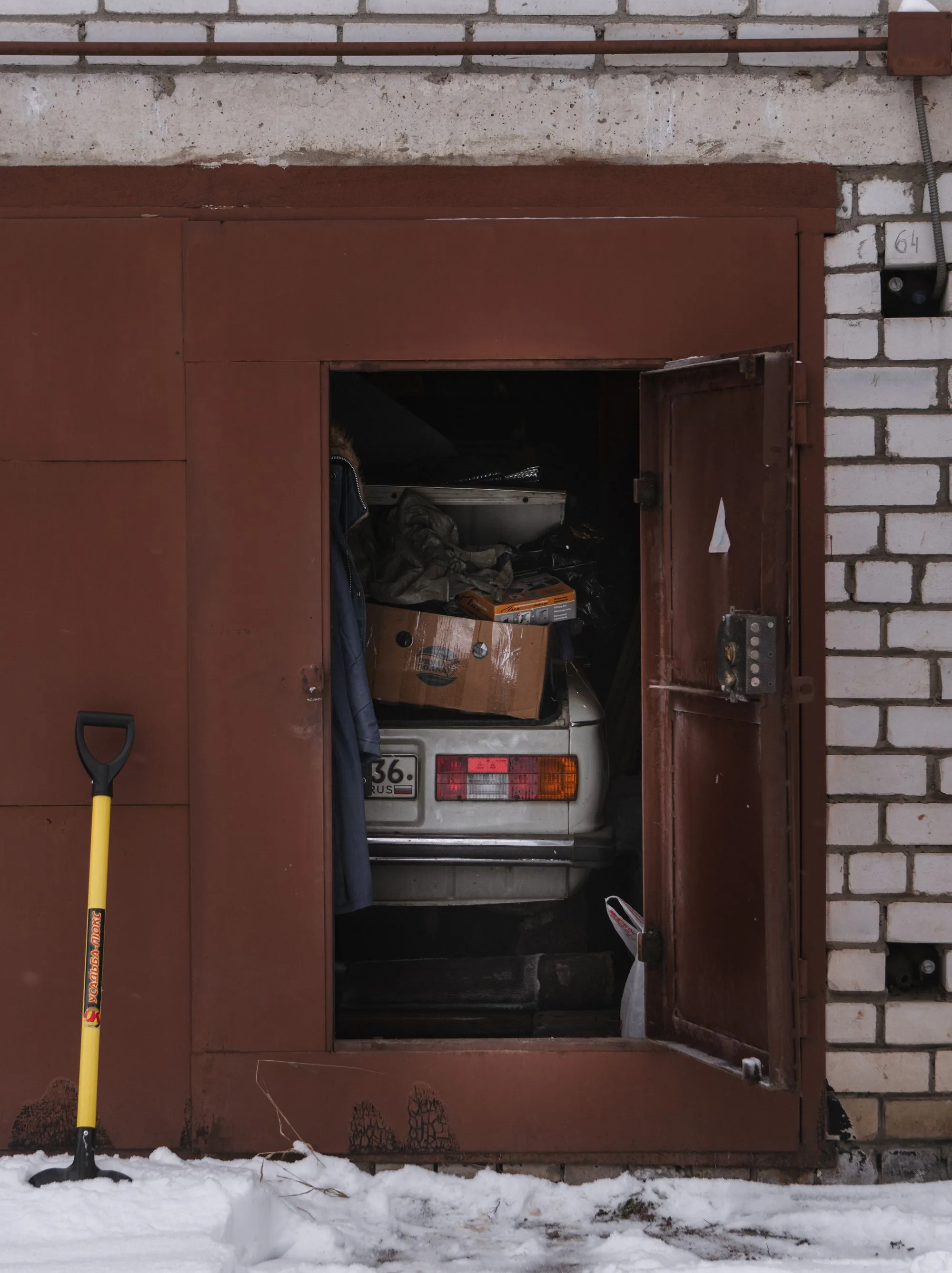 An old white car parked inside a narrow brick garage with the door open and snow on the ground.