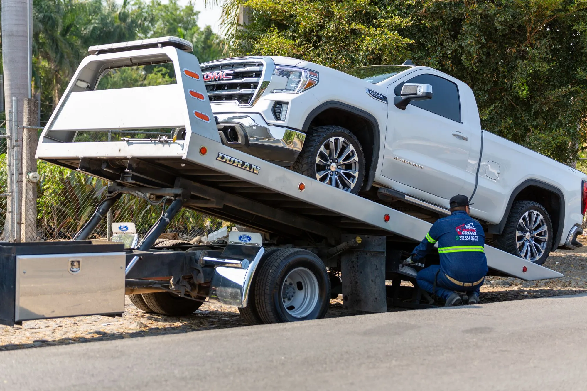 A tow operator chains a white pickup truck onto a tilted flatbed at the curb.