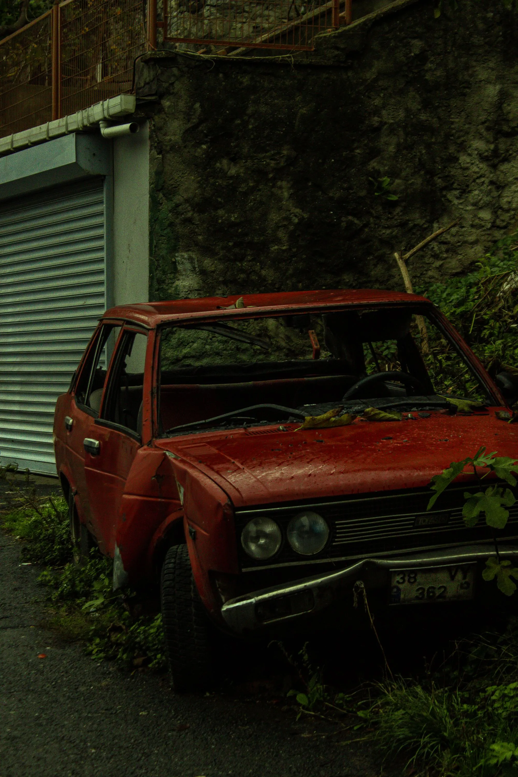 A faded red sedan with flat tires sits among weeds in a narrow alley.