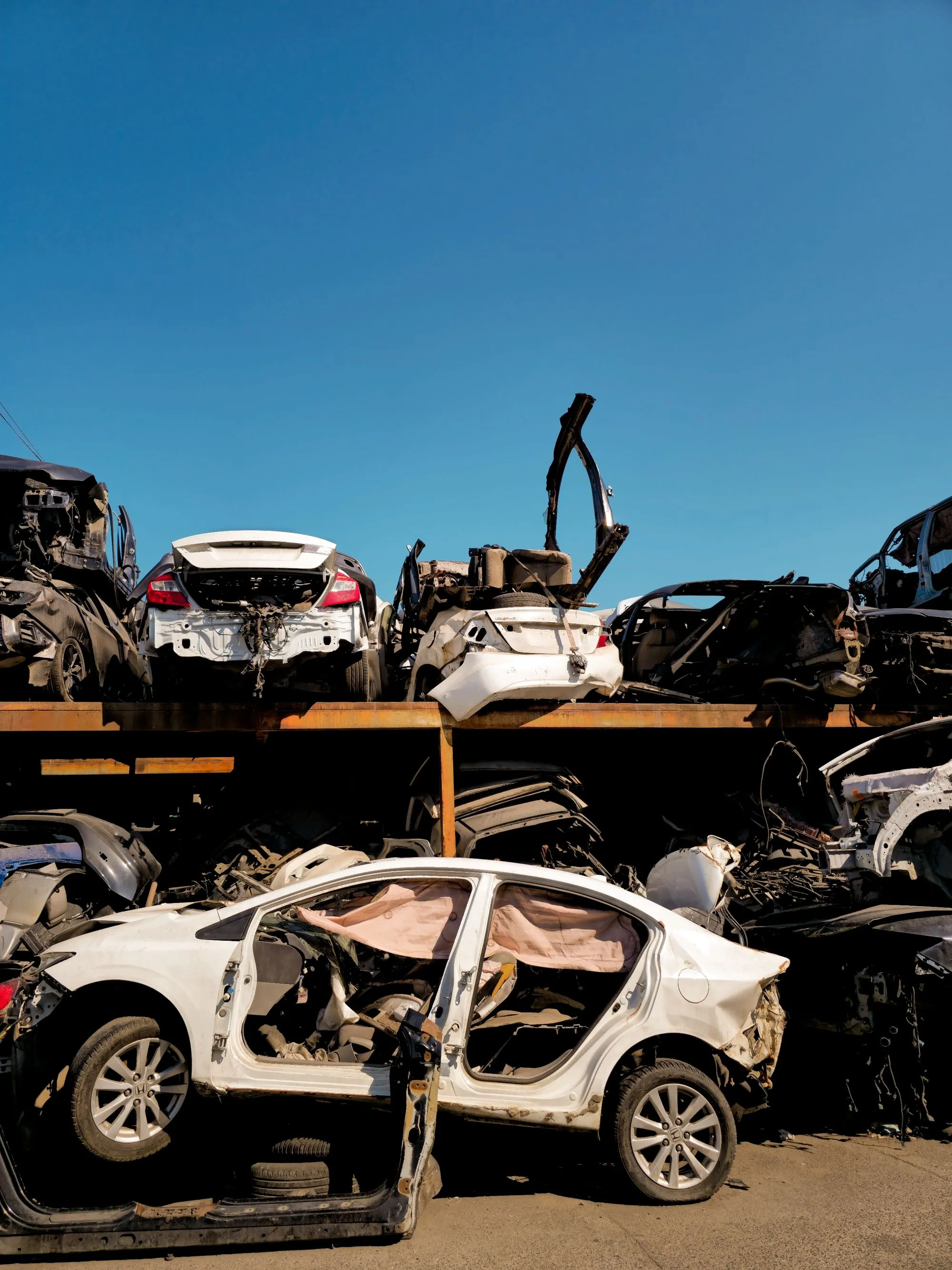 Rows of stripped and crushed cars stacked on racks at a scrapyard under a blue sky.