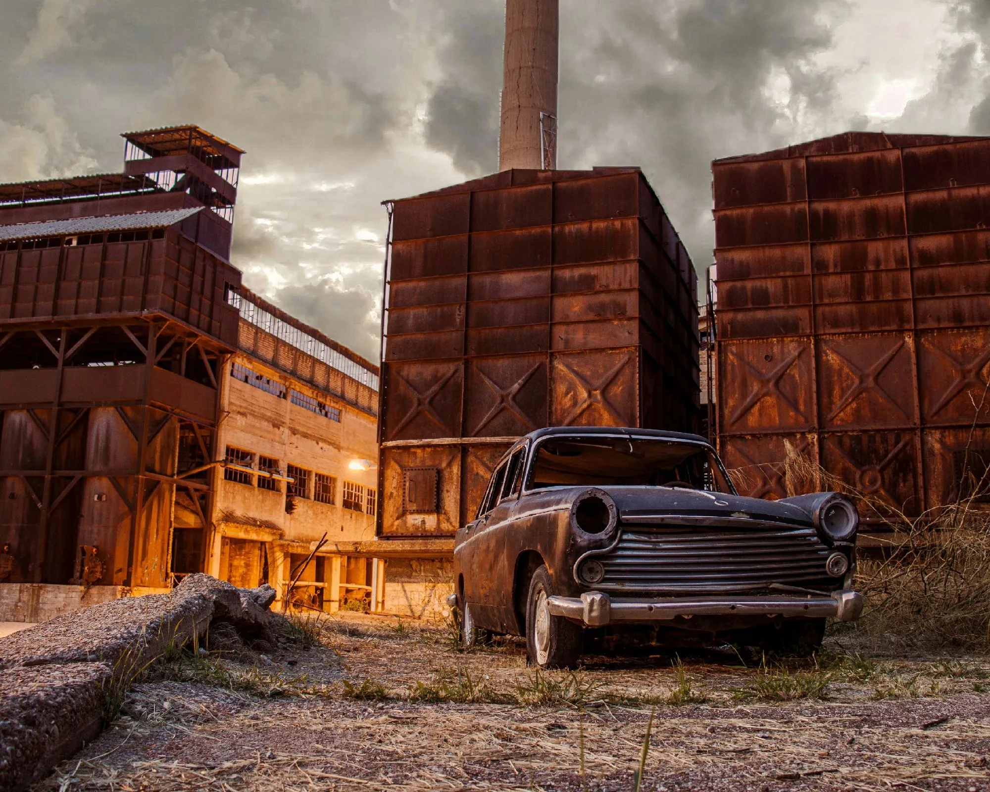 A rusty old sedan parked in front of a weathered brick factory under a cloudy sky.
